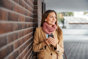 Mobile networks and providers. A happy young woman in a coat leaning on the brick wall outdoors, holding a smart phone, and looking away.