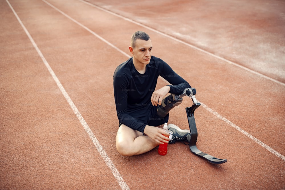Un corredor cansado con una pierna ortopédica sentado en la pista de atletismo con un refresco en la mano y tomando un descanso.