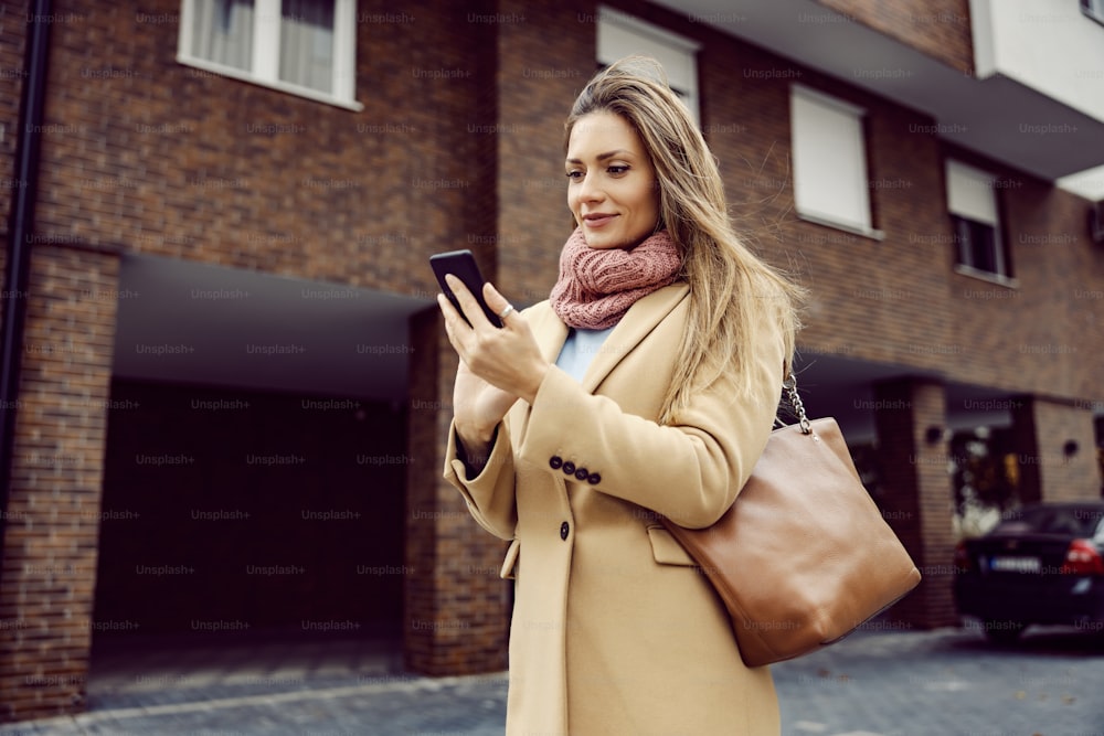 Wifi network, global communications, and mobile providers. A young woman in warm clothes standing on the street in cold windy weather and typing on her mobile phone.