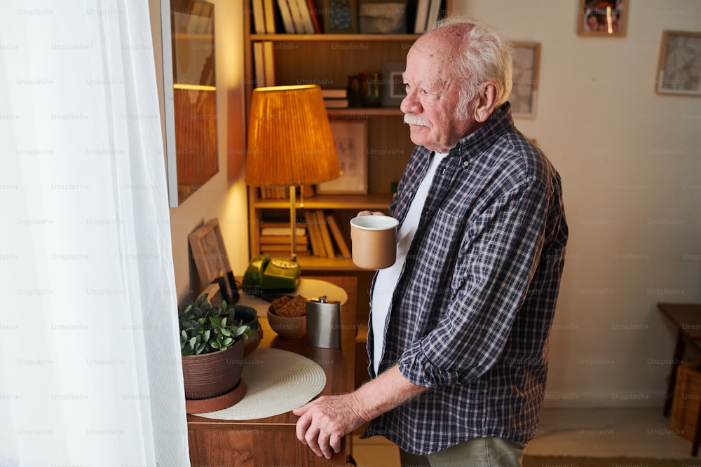 朝、白いナイロンカーテンの窓の前に立ち、外を眺めるお茶の入ったマグカップを持った年配の男性
