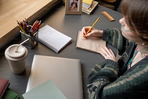 Adolescente criativo com lápis sobre a página do bloco de notas sentado ao lado da mesa e pensando em novas ideias enquanto desenha à vontade