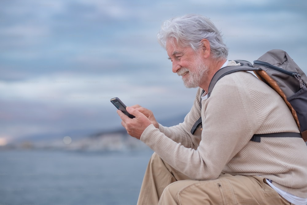 Smiling adult mature old senior man sitting on the cliff in sea excursion using mobile phone. Caucasian white haired grandfather enjoying good time, technology and social media during retirement