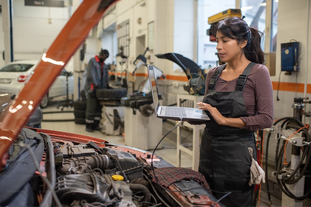 Retrato de vista lateral de una joven mecánica usando una computadora portátil mientras inspecciona el vehículo en el taller de reparación de automóviles, espacio de copia