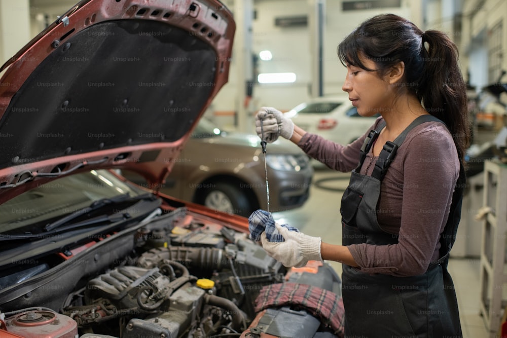 Retrato da vista lateral da mecânica feminina jovem verificando os níveis de óleo enquanto conserta o carro na oficina, espaço de cópia