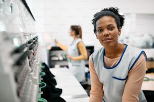 Jeune couturière afro-américaine travaillant à la machine à broder dans une usine textile et regardant la caméra.