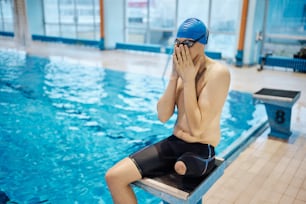 Male swimmer without one leg sitting on starting block and adjusting his swimming goggles before getting into the water.