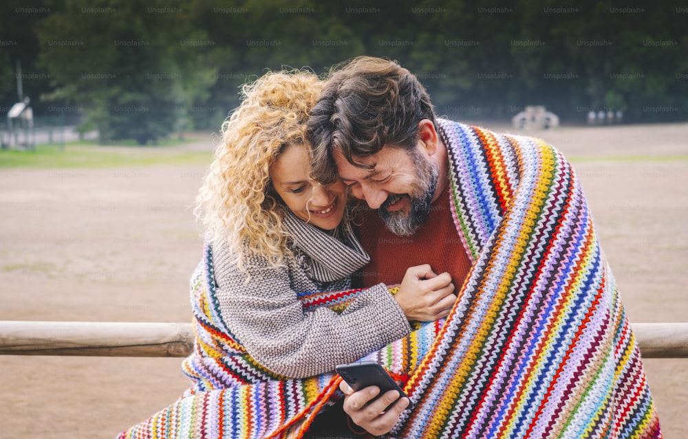 Adult caucasian couple enjoy love and relationship using together a smart phone device and having fun covered with colorful knit wool. Happy man and woman outdoor with cell connection