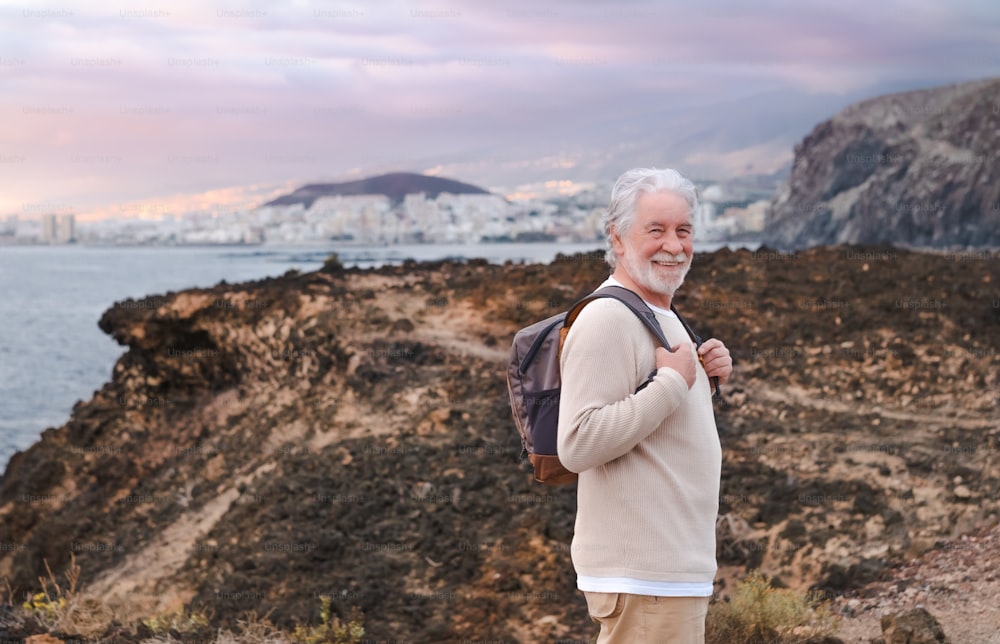 Adult caucasian gray haired senior man walking in countryside close to the sea wearing backpack looking at camera smiling enjoying nature. Freedom and vacation concept. Cloudy sky. Copy space
