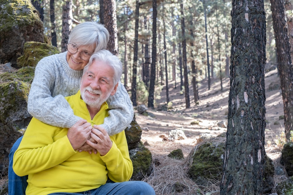 手をつないで森に座っている美しい幸せな老夫婦。笑顔で夫を抱きしめる白人女性。晴れた日に休暇の自由と自然ハイキングを楽しむ年配の白髪の人々