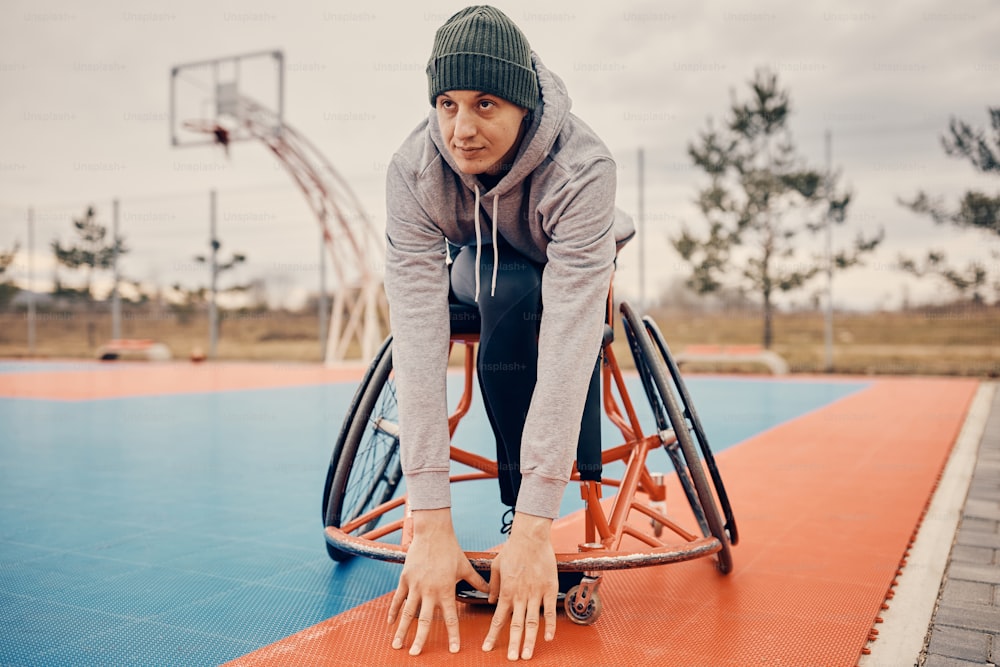 Joven jugador de baloncesto en silla de ruedas haciendo ejercicios de estiramiento en la cancha al aire libre antes del entrenamiento deportivo.