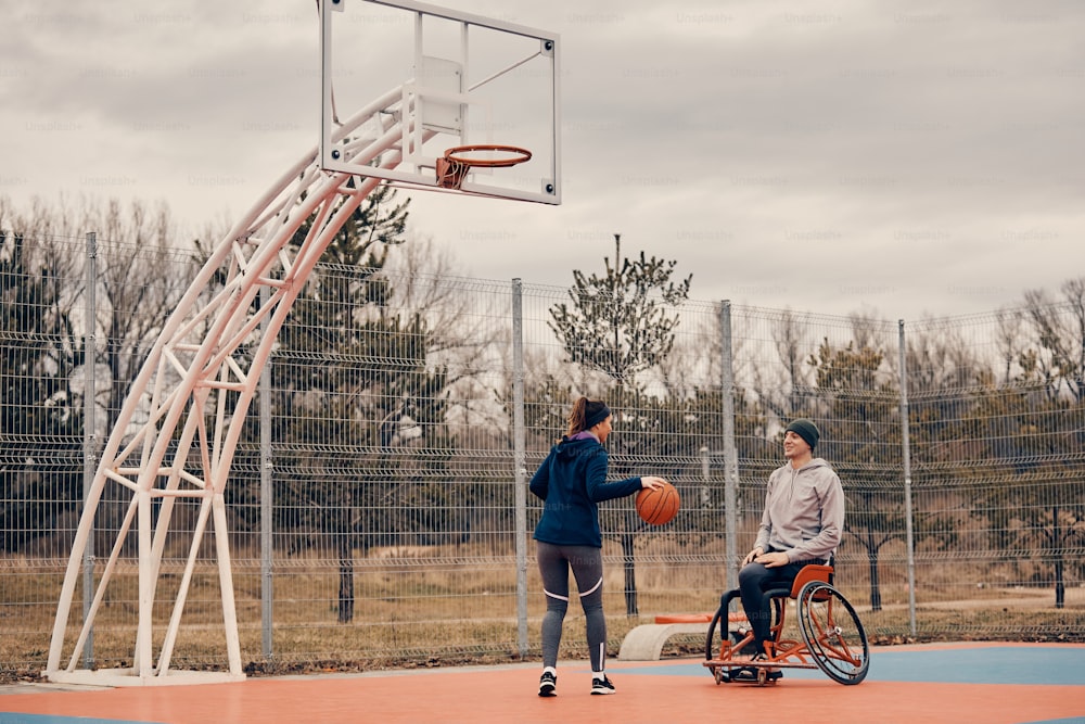 Deportista feliz con discapacidad usa silla de ruedas mientras juega baloncesto con una amiga al aire libre. Espacio de copia.