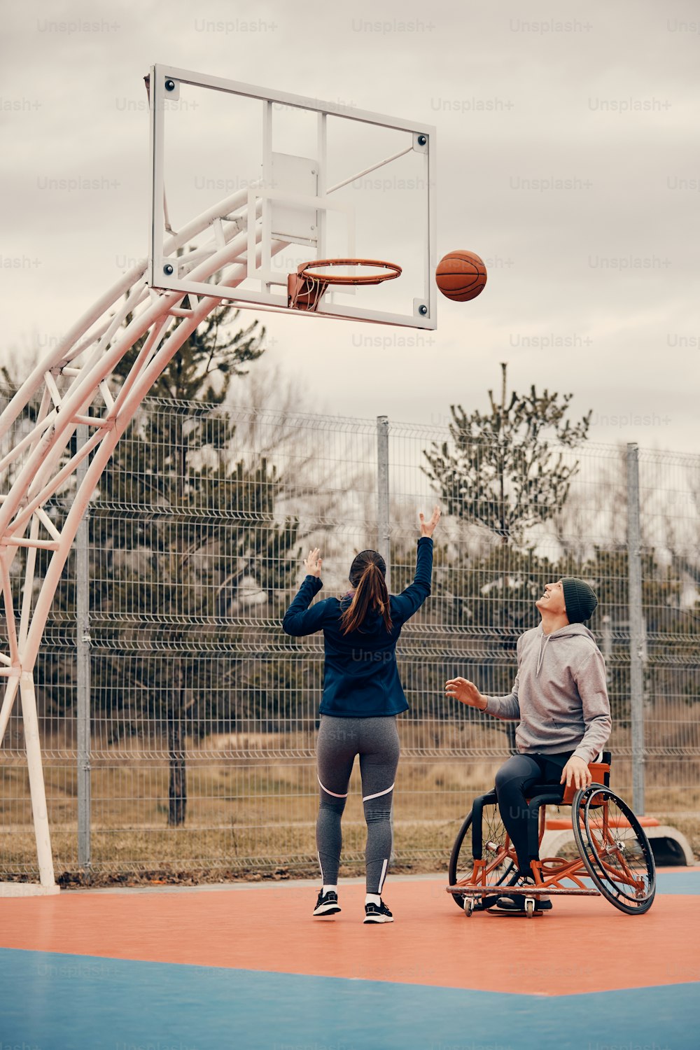 Rear view of athletic woman shooting at the hoop while playing basketball with a friend who uses wheelchair.