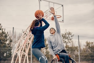 Happy sportsman with disability playing basketball with female friend and trying to block her while she is shooting at the hoop.