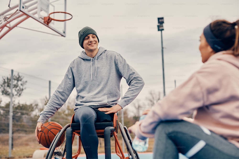 Happy athlete in wheelchair communicating with female friend while relaxing after playing basketball on outdoor sports court.