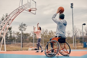Rear view of man with disability playing basketball with his girlfriend and shooting at the hoop outdoors.