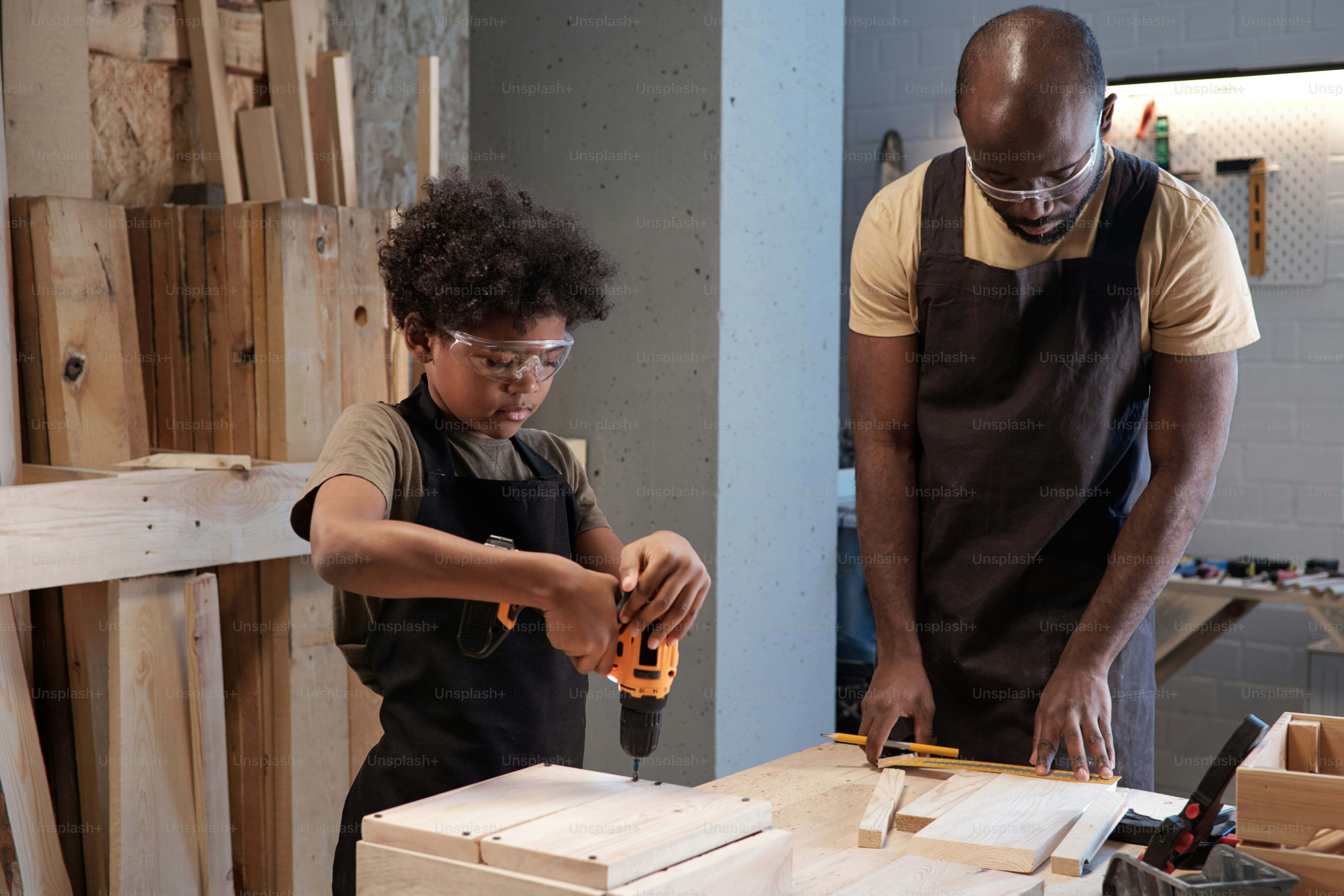 Side view portrait of cute African-American boy building wooden birdhouse and using power drill in workshop with father