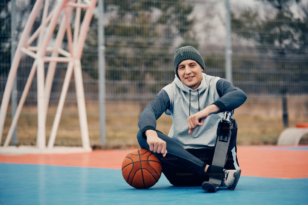 Young athlete with prosthetic leg relaxing on outdoor basketball court and looking at camera.