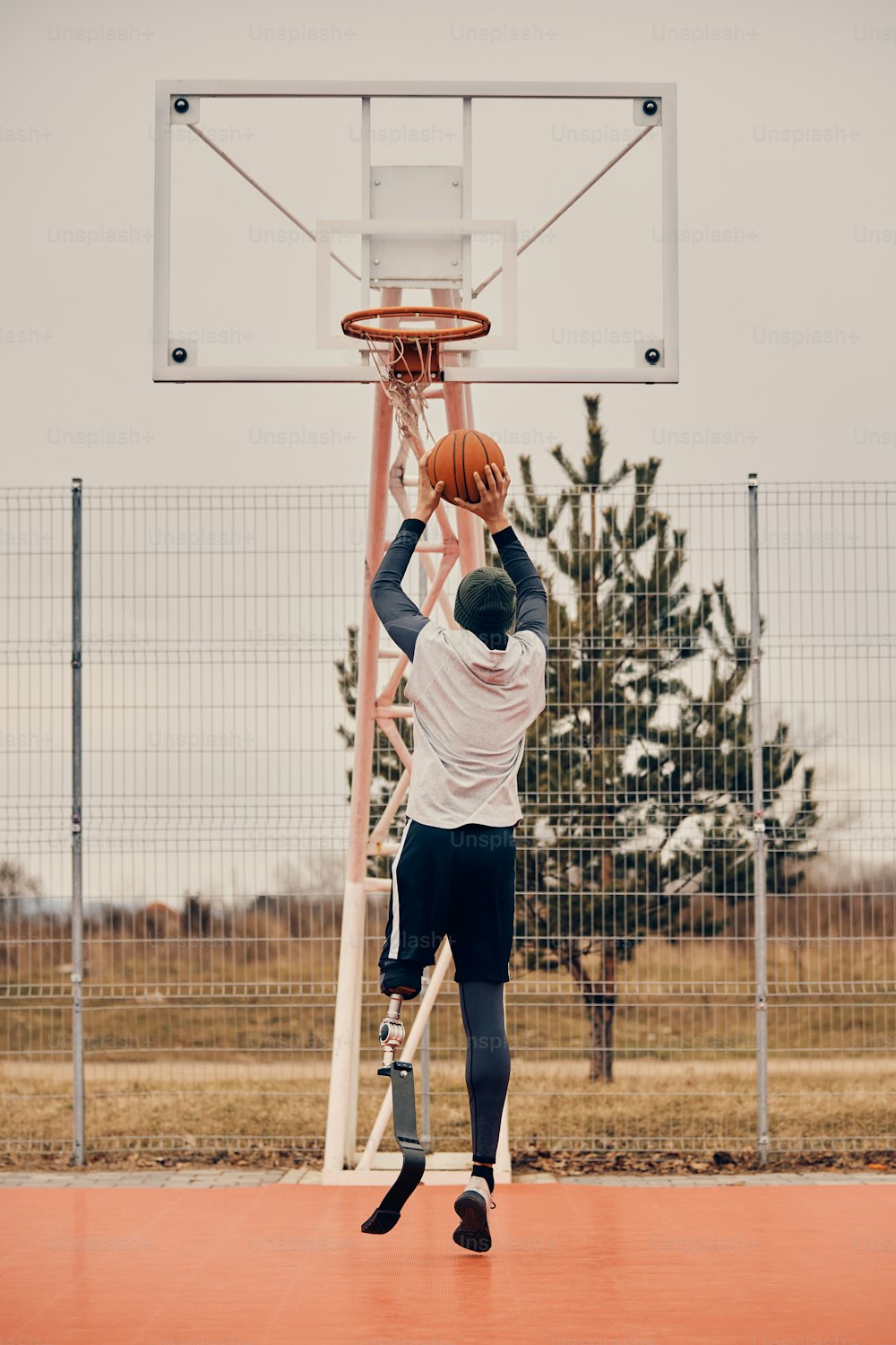 Vista trasera de un jugador de baloncesto con una pierna protésica tomando un tiro mientras practica en una cancha deportiva al aire libre.