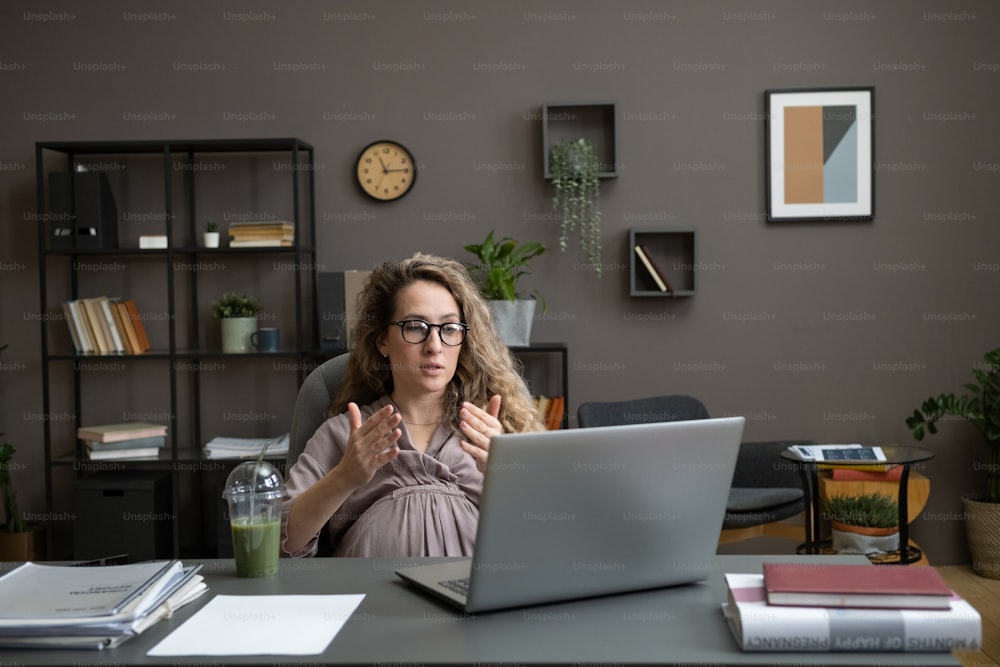 Giovane donna d'affari incinta che si siede sulla sedia al tavolo e che ha una conversazione online sul computer portatile durante il lavoro in ufficio