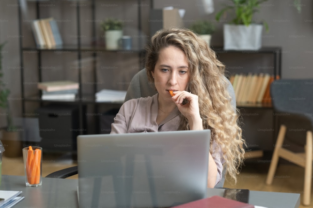 Joven mujer de negocios con cabello largo y rizado comiendo zanahoria y mirando el monitor de la computadora portátil mientras está sentada en la mesa de la oficina