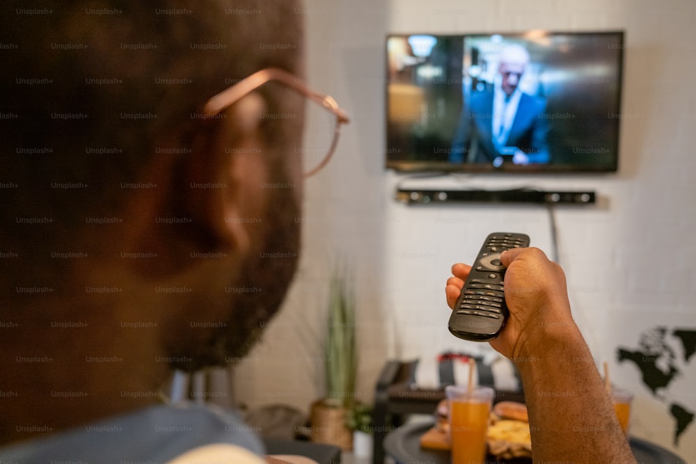 Nahaufnahme eines afrikanischen Mannes mit Brille, der in seiner Freizeit zu Hause mit Fernbedienung die Sender im Fernsehen umschaltet