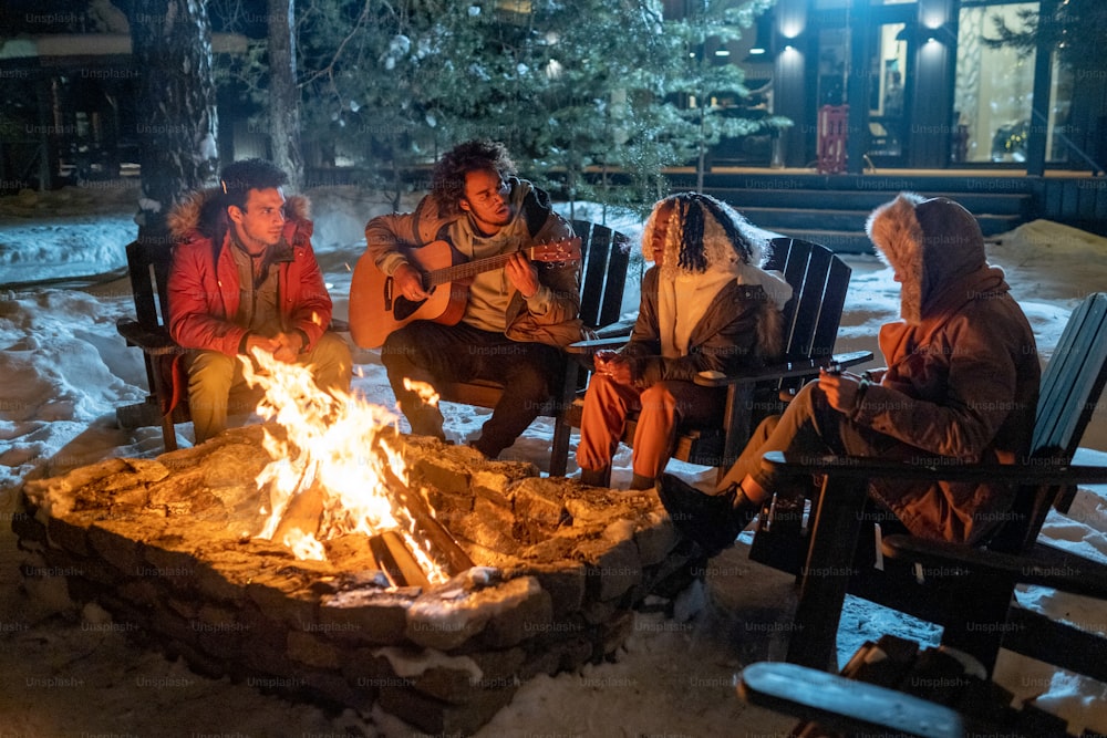 Groupe d’amis jouant de la guitare et chantant des chansons ensemble tout en étant assis près du feu à l’extérieur un soir d’hiver