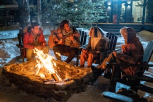 Grupo de amigos tocando la guitarra y cantando canciones juntos mientras están sentados cerca del fuego al aire libre en la noche de invierno