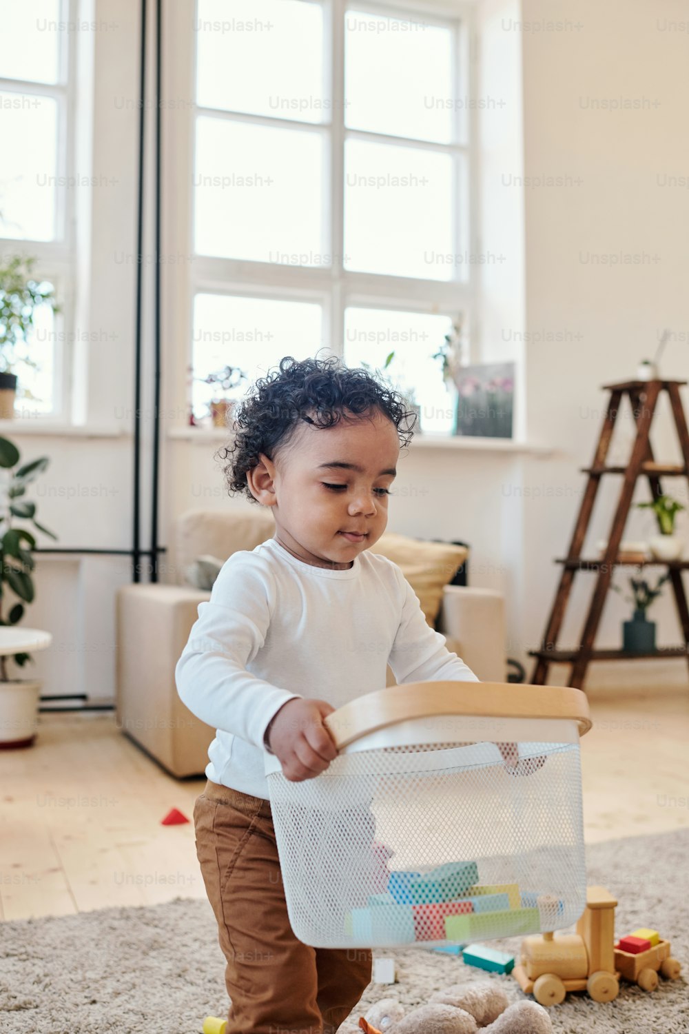 Cute little Black boy with curly hair walking across living room and picking up toys into container