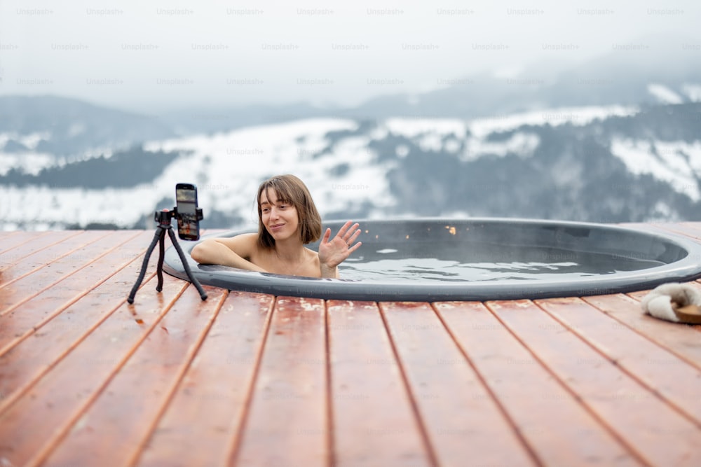 Donna che blogga se stessa al telefono mentre nuota in una vasca calda in montagna. Concetto di influencer e riposo sulla natura. Idea di evasione e svago in montagna