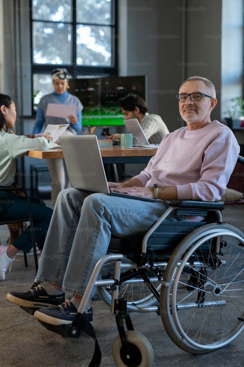 Mature employee with laptop on his knees sitting in wheelchair in front of camera against group of young intercultural colleagues
