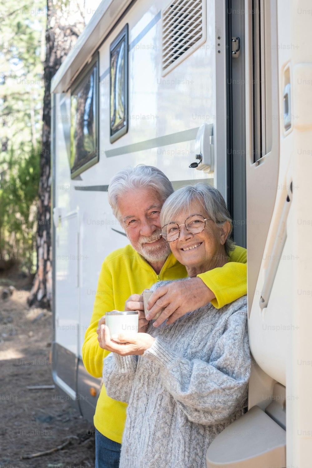 Happy relaxed caucasian senior couple leaning outdoor at their camper van motor home holding coffee cups. Smiling attractive elderly people enjoying freedom vacation travel in the forest.