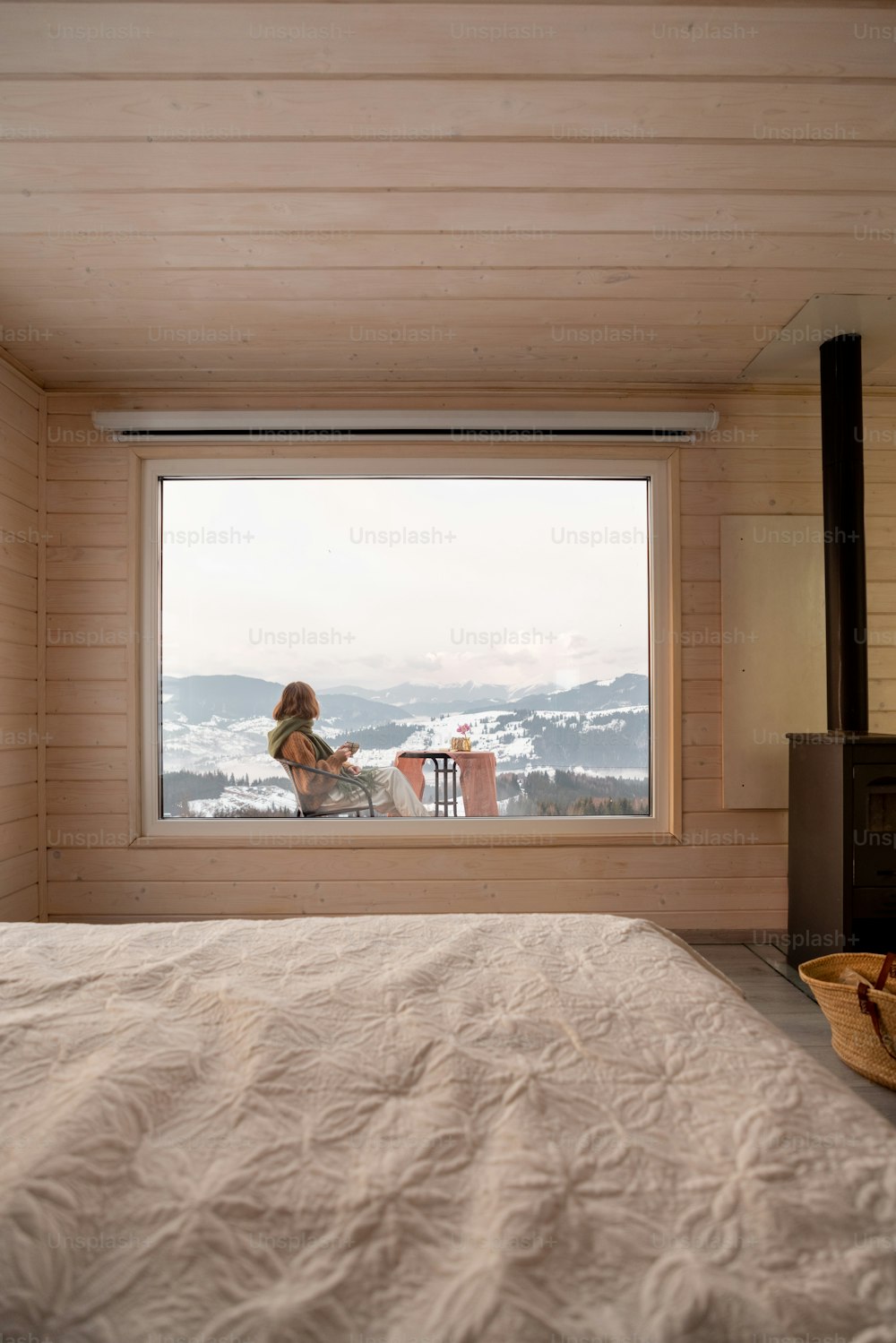 Woman sitting with a drink on terrace of tiny house in the mountains, view through the window from the inside. Concept of small modern cabins for rest and escape to nature