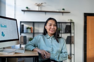 Young Asian female economist in smart casualwear standing by workplace with computer monitor and financial data on screen