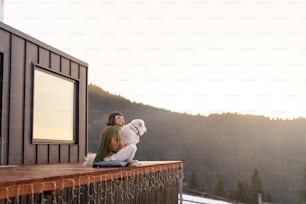 Frau sitzt mit Hund auf der Terrasse eines winzigen Hauses in den Bergen. Konzept der kleinen modernen Kabinen für Ruhe und Flucht in die Natur. Idee, mit Hund zu reisen