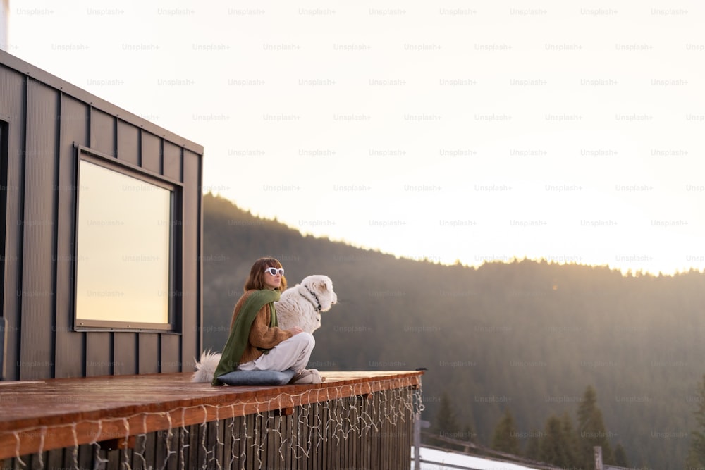 Woman sitting with dog on terrace of tiny house in the mountains. Concept of small modern cabins for rest and escape to nature. Idea of traveling with dog