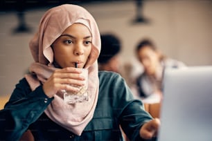 Junge muslimische Frau, die Kaffee trinkt, während sie auf einem Laptop in einem Café im Internet surft,