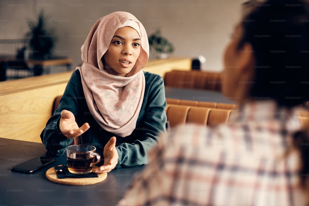 Young Islamic woman in hijab communicating with female friend while drinking tea with her in a cafe.