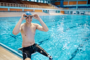 Happy athlete with a leg disability practicing swimming at indoor pool and looking at camera.