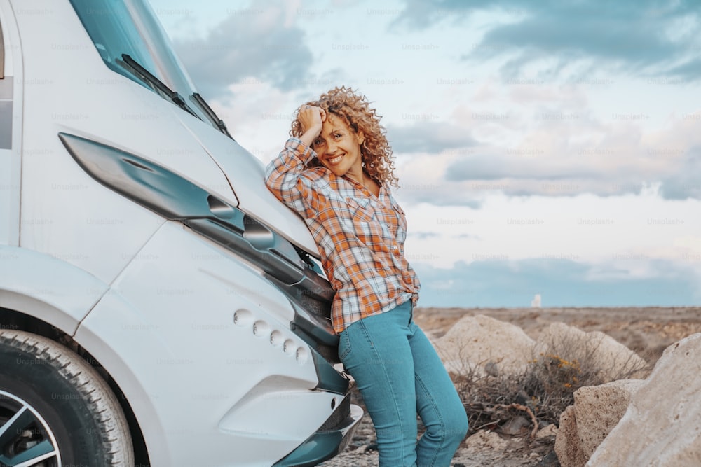Happy adult woman tourist against modern camper van motorhome smile and look at the camera. Female people traveler enjoy destination and vacation. Country side and blue sky in background