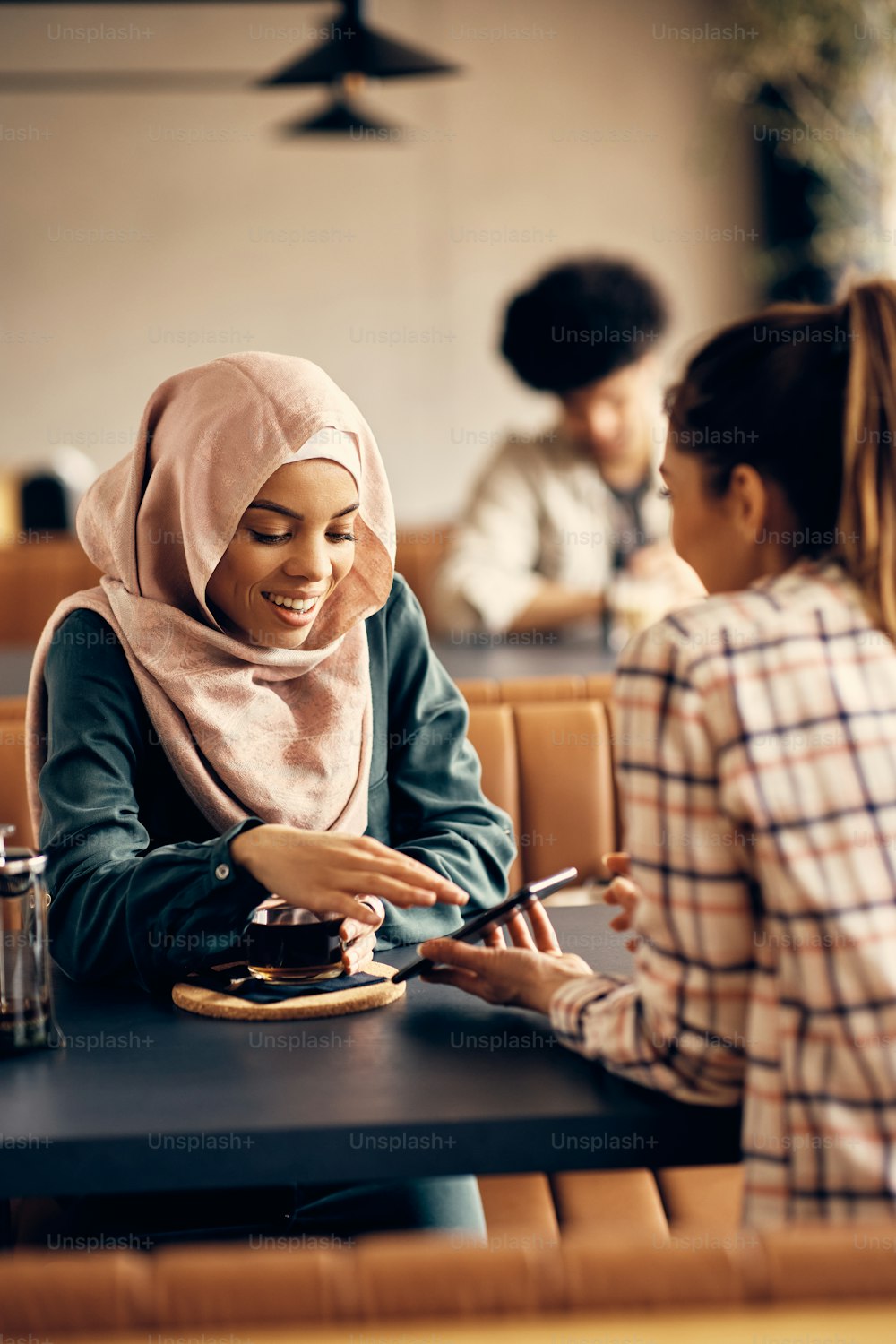 Happy Islamic woman reading text message on her friend's cell phone in a cafe.
