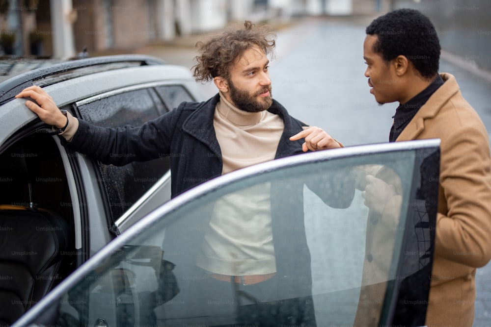 Two men flirting or having a close conversation while opening car door on the street. Concept of homosexual relations or close male friendship. Caucasian and hispanic man wearing coats