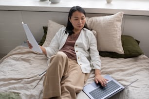 Young serious female auditor concentrating on work with documents and online financial data on laptop screen while sitting on bed