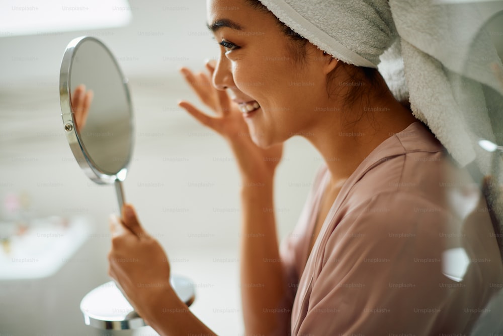 Happy Asian woman applying face cream while looking herself in a mirror in the bathroom.