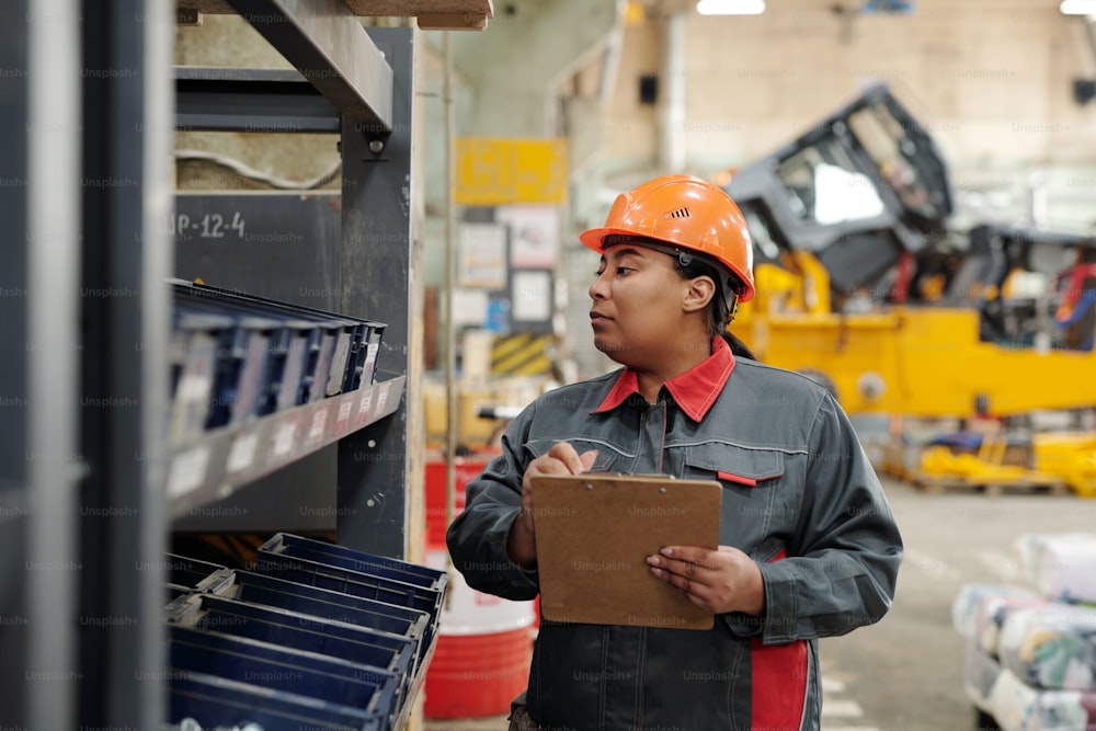 Female worker of industrial plant making revision notes in document while looking through several plastic containers with spare parts