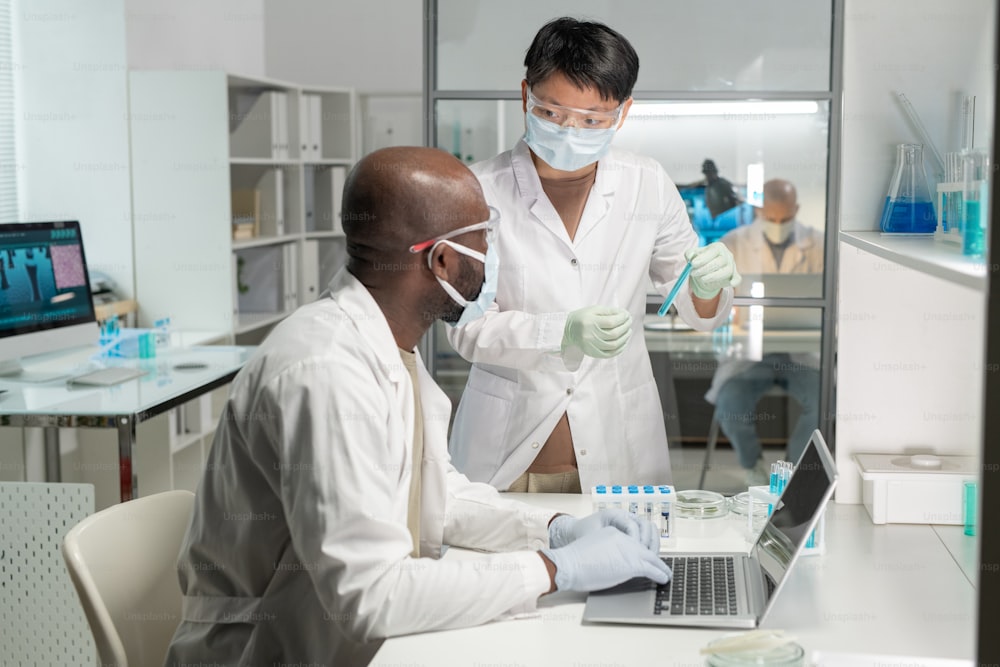 Joven científica asiática con máscara protectora y bata de laboratorio que muestra a su colega afroamericano frasco con líquido azul