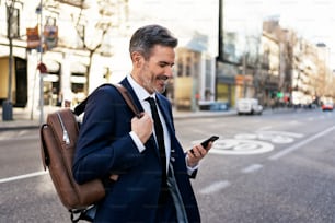 Side view of content mature male entrepreneur in suit and with backpack crossing road and reading message on mobile phone