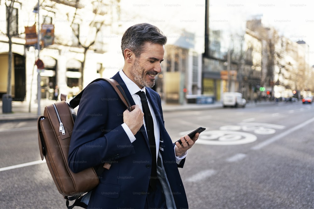 Vista laterale del contenuto imprenditore maschio maturo in giacca e cravatta e con zaino che attraversa la strada e legge il messaggio sul telefono cellulare