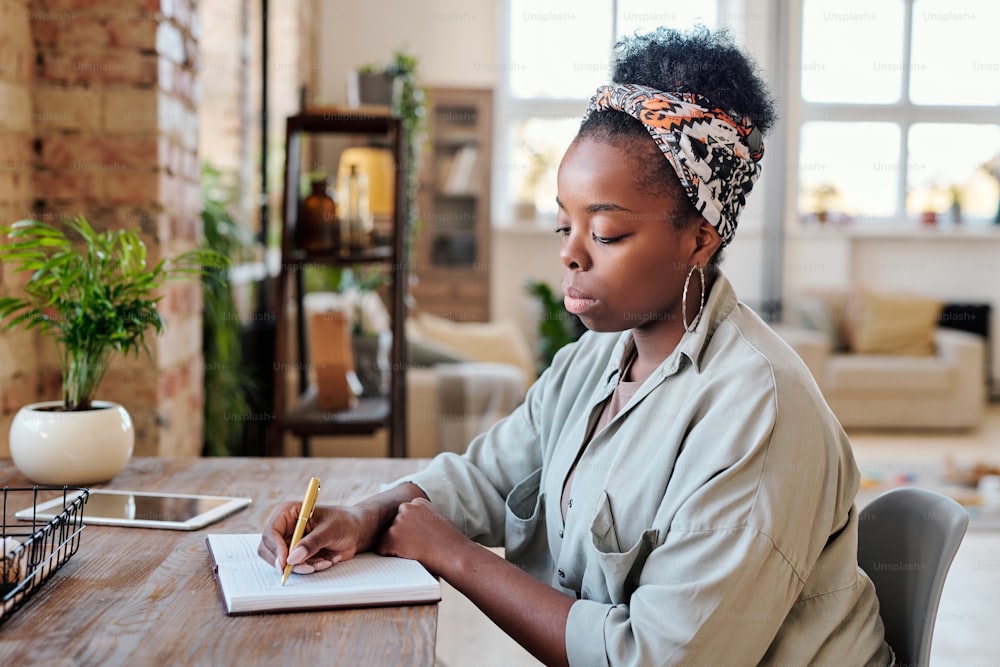 Serious young Black woman in earrings sitting at wooden desk and making notes in planner