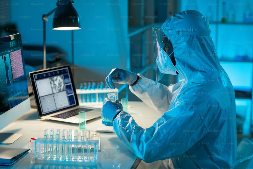 Young African American male scientist in protective coveralls studying liquid substance while sitting by desk in laboratory