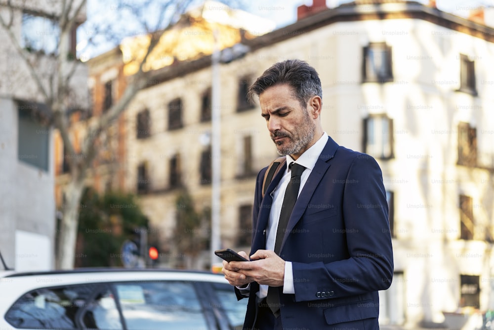 Focused male entrepreneur in elegant suit messaging on mobile phone while walking along street